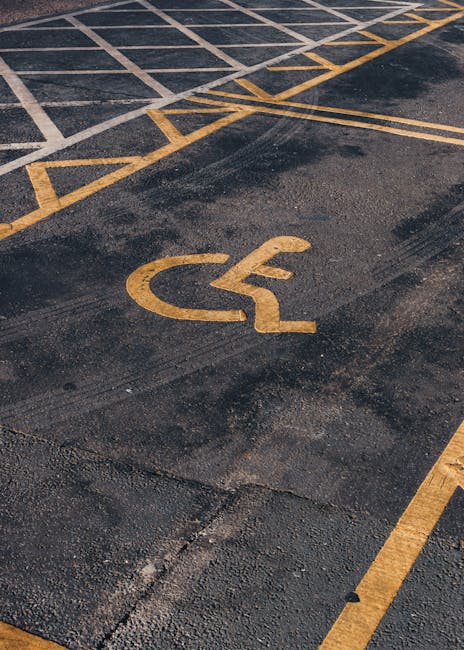 A close-up view of an accessible parking space with a yellow-painted wheelchair symbol on dark asphalt. The space is marked with yellow lines and diagonal hatching in the background, indicating a desi