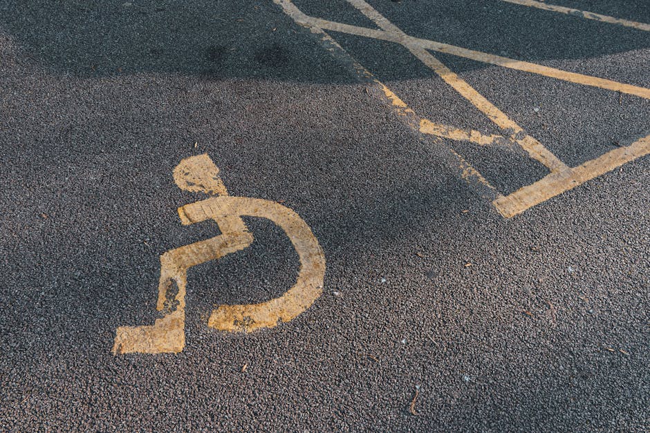 A close-up view of an accessible parking space with a yellow-painted wheelchair symbol on dark asphalt. The space is marked with yellow lines and diagonal hatching in the background, indicating a desi